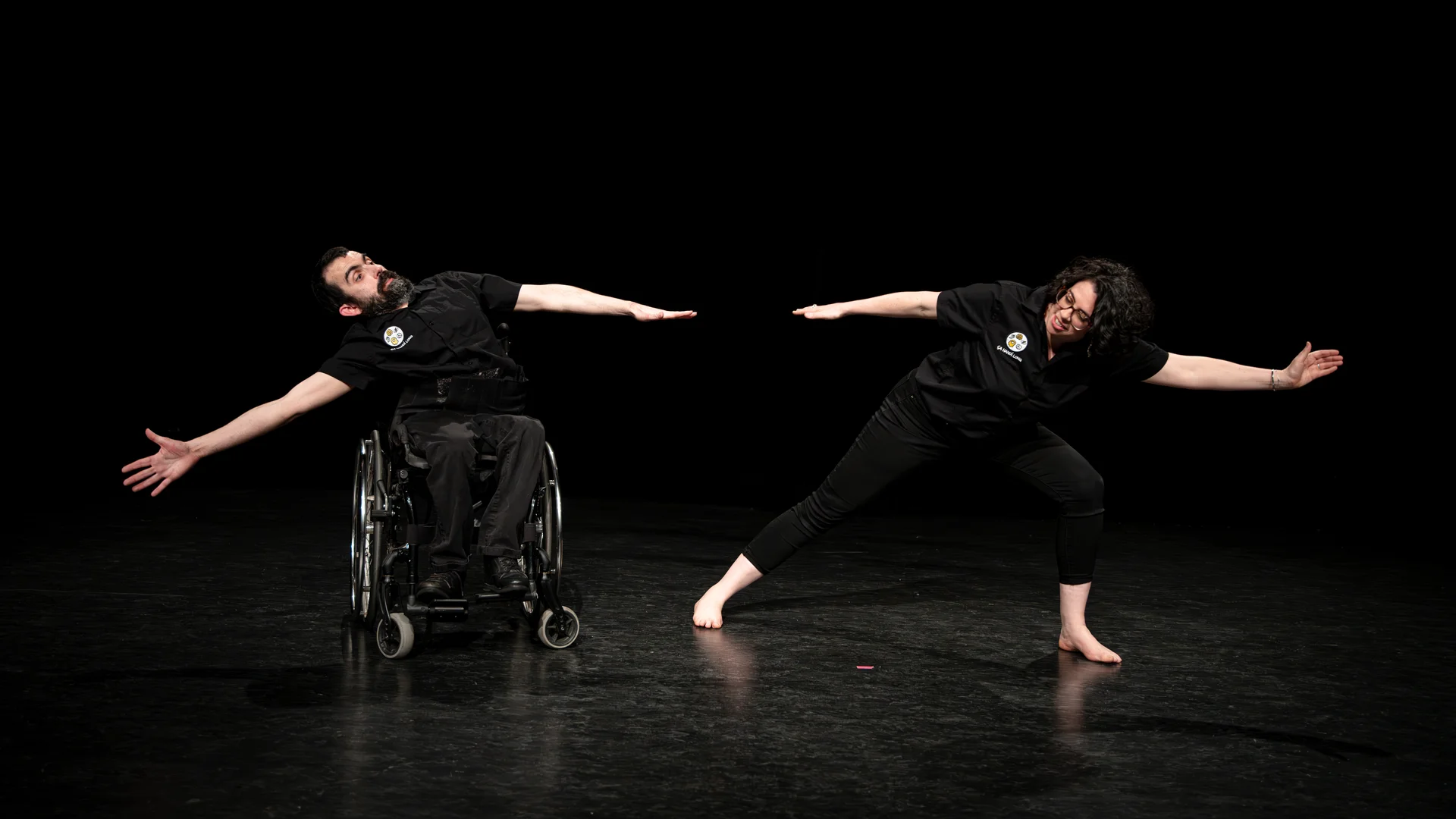 Scène du spectacle Ça handi long. Simon Garnes et Clémence Fouquet sont côte à côte et de face. Simon tend ses bras de chaque côté de son corps en se penchant vers la gauche de la photo, il est en fauteuil roulant. Clémence est sur ses jambes, dans un mouvement de fente vers la droite et tend aussi ses bras de chaque côté de son corps. Les deux sont en chemises noires avec le logo Ça handi long et en pantalon noir.