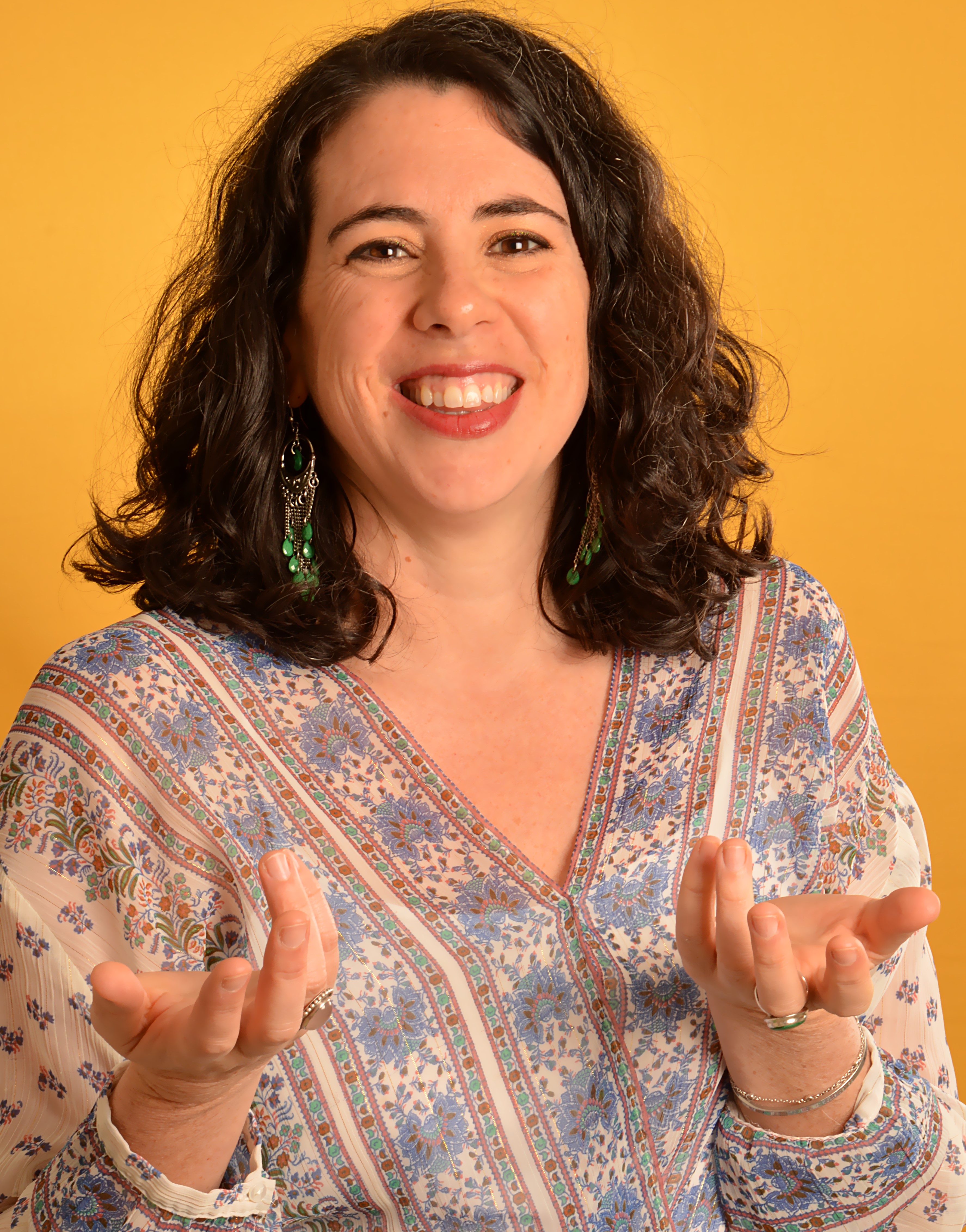 Clémence Fouquet - Consultante RH Diversité et Inclusion, portrait de face de la tête à la taille, Clémence Fouquet sourit, tend les deux mains vers l'avant de la photo, elle porte un haut blanc avec des lignes fines roses et dorées avec des motifs floraux bleus et dorés entre